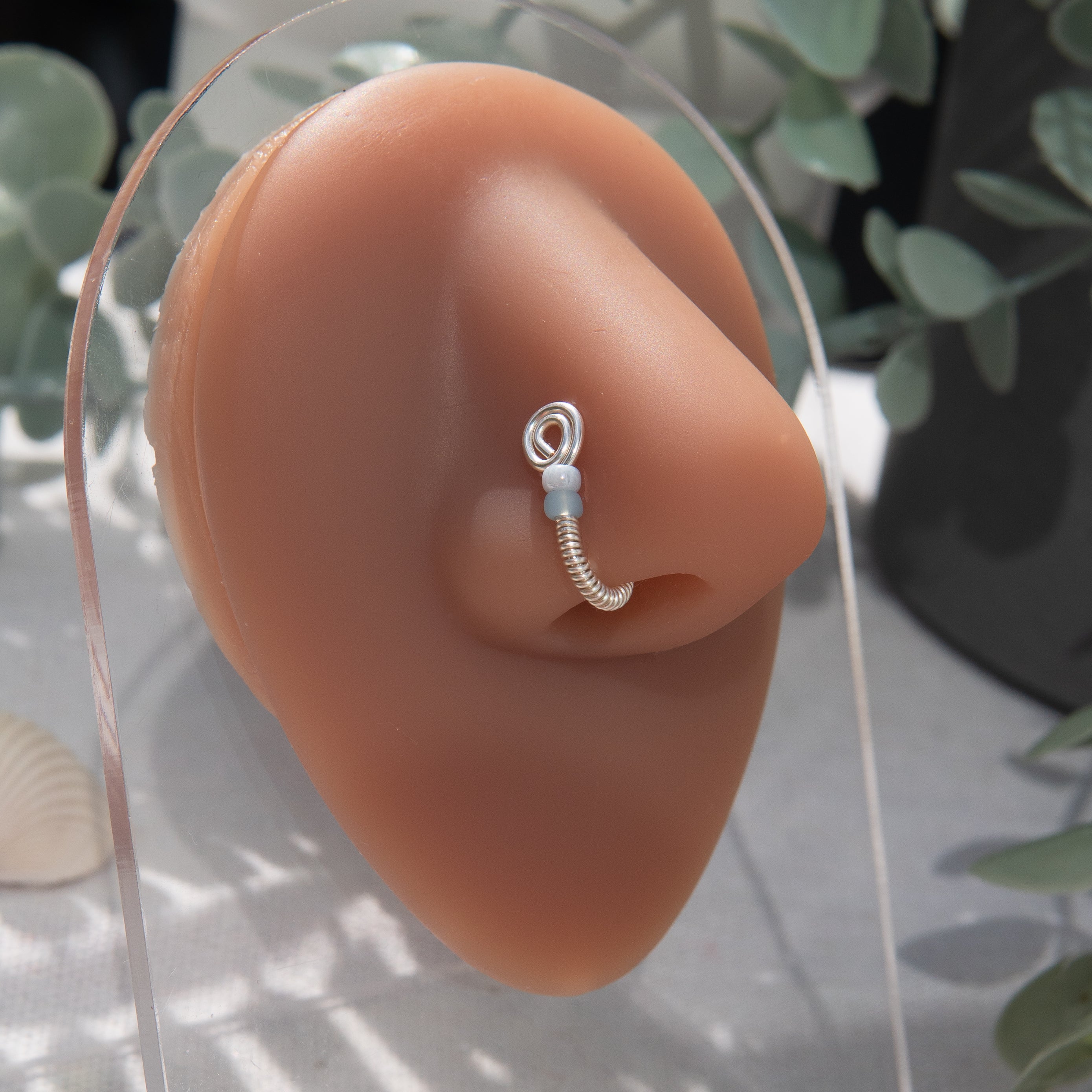 A silver-toned fake nose ring cuffs the model's nostril. The wire design forms a circular shape. Two seed beads rest below the circle. 