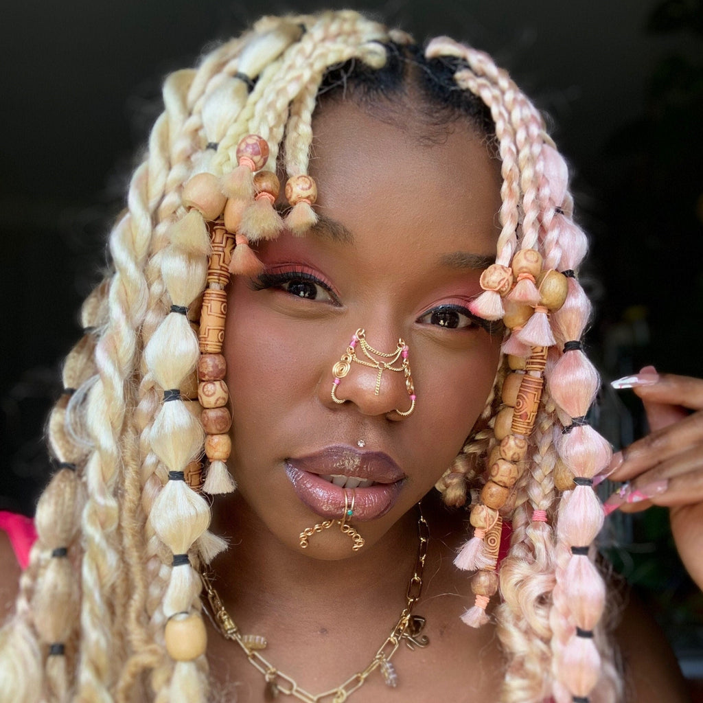 A gold-toned faux chain bridge nose piercing cuffs both of the model's nostrils. Two dainty 18K gold-plated curb chains rest across the bridge of the nose attached to the top and middle of each cuff, forming a criss-cross shape. Three pink seed beads were interweaved into the design of each nose cuff.