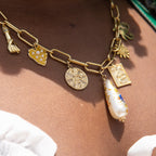 A chunky gold-plated charm necklace made with a chunky 18K gold-plated curb chain. Multiple gold-plated charms hang along the length of the necklace in the following order: seashell, padlock, scarab beetle, a green heart, crescent moon, Duafe symbol, circle disc with the engravement 'thankful, grateful, blessed'