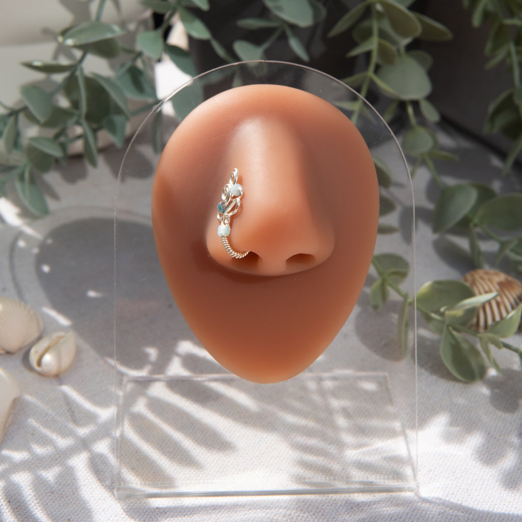 A silver fake nose piercing cuffs the model's nostril. The wire design forms an abstract spiral shape with three blue seed beads intertwined into the design.