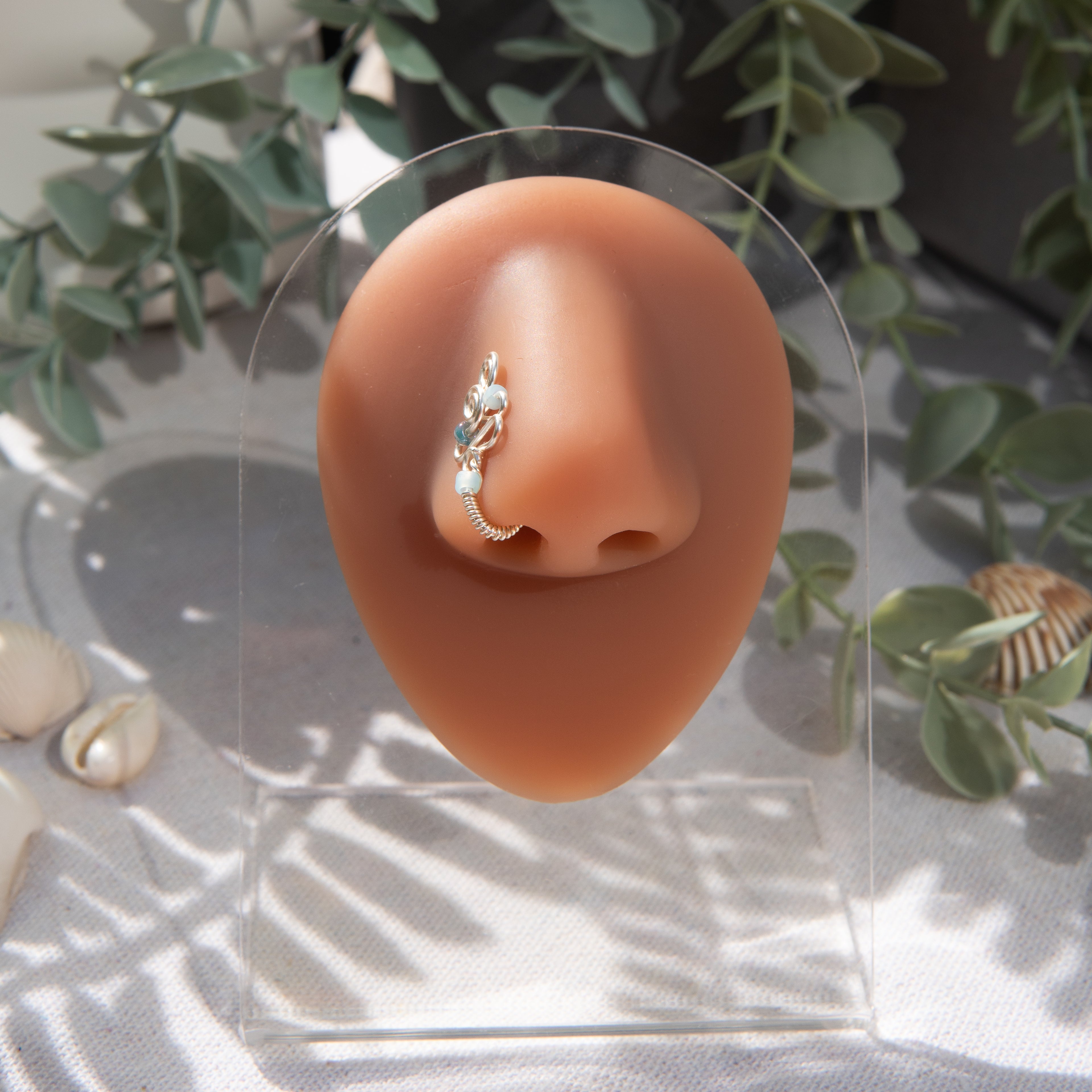 A silver fake nose piercing cuffs the model's nostril. The wire design forms an abstract spiral shape with three blue seed beads intertwined into the design.