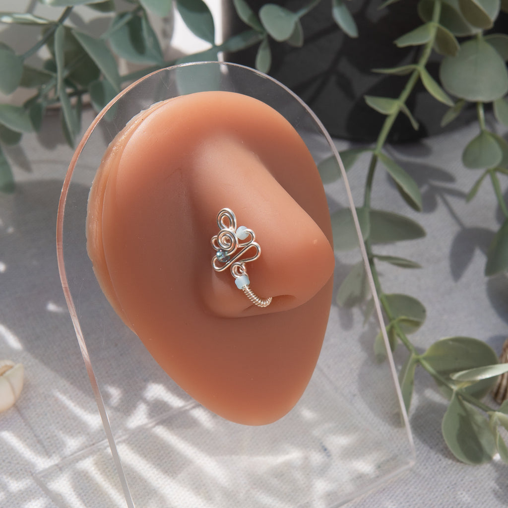 A silver fake nose piercing cuffs the model's nostril. The wire design forms an abstract spiral shape with three blue seed beads intertwined into the design.