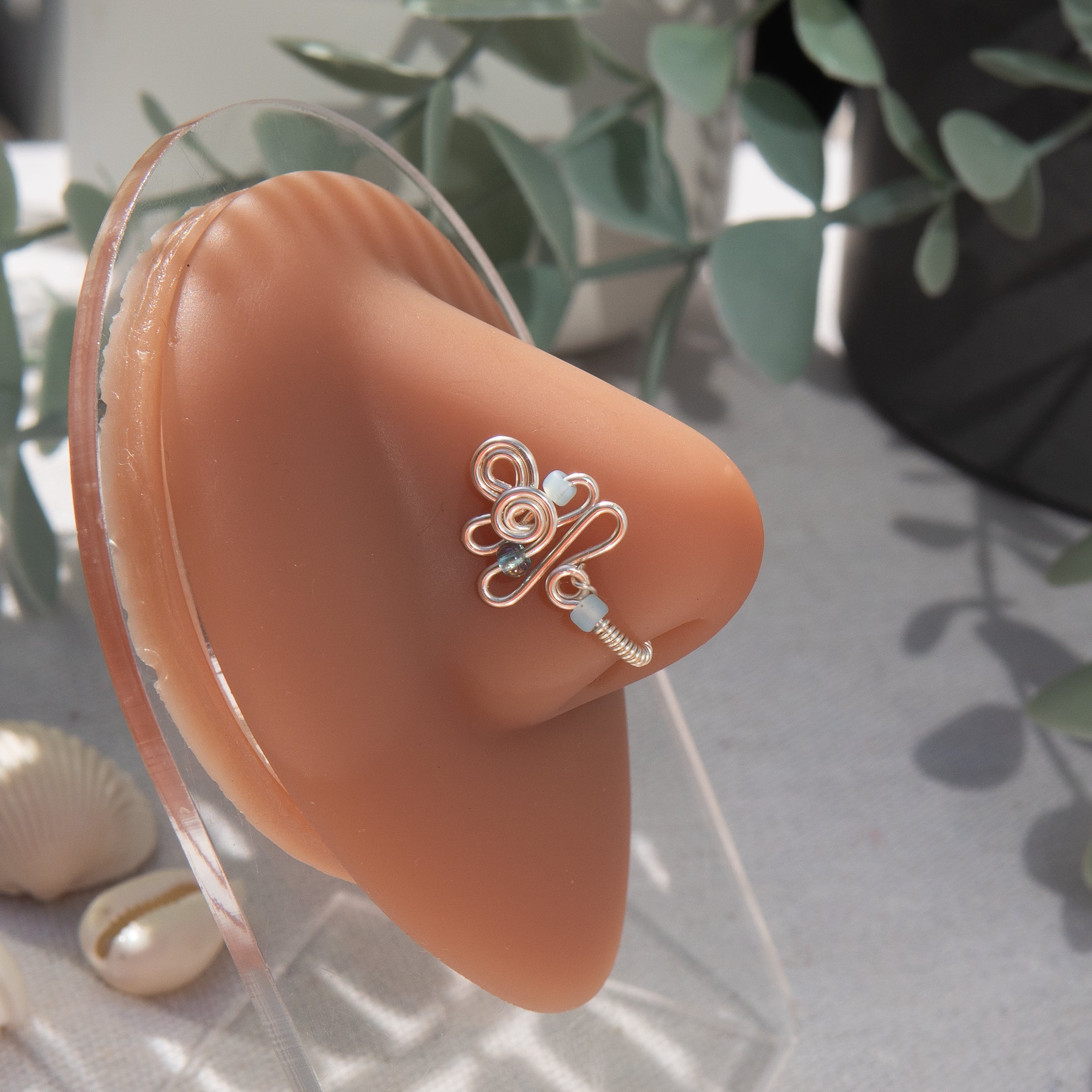 A silver fake nose piercing cuffs the model's nostril. The wire design forms an abstract spiral shape with three blue seed beads intertwined into the design.